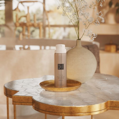 A beige spray bottle on a round marble table with gold edges, next to a large beige vase with white flowers.