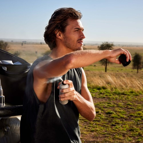 A man sprays sunscreen on his arm outdoors in a field, with a backpack and trees in the background.