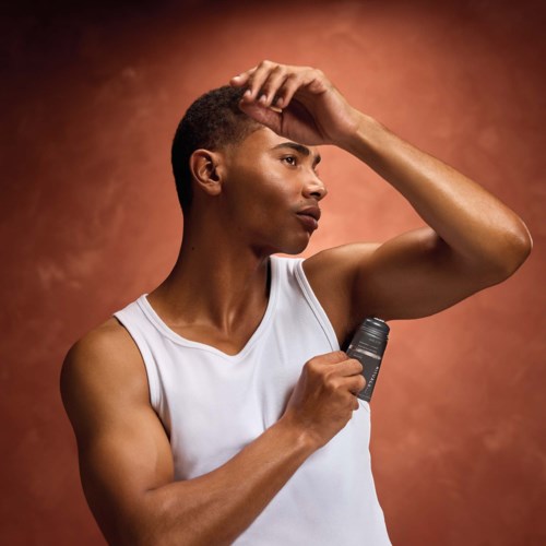 A man with short hair and a white sleeveless shirt, holding a deodorant, looking thoughtful, against a brown background.