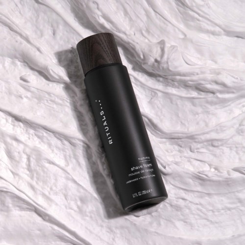 A black cylindrical container with a brown cap lying on white crumpled fabric, labeled RITUALS, shave foam.