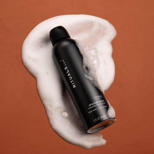 Black bottle of Rituals shower foam lying on soap bubbles on a brown surface.