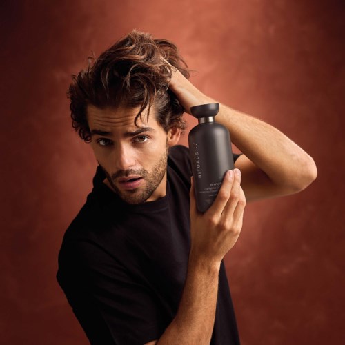 A man with messy hair holding a matte black bottle of hair care product against a brown background.