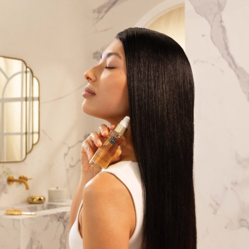 A woman with long, straight black hair sprays her hair in a bathroom with marble walls and golden fixtures.