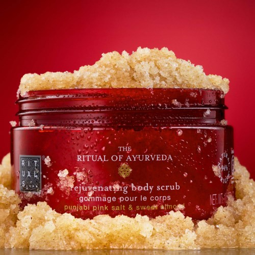 A red jar titled The Ritual of Ayurveda with body scrub and sand around it on a red background.