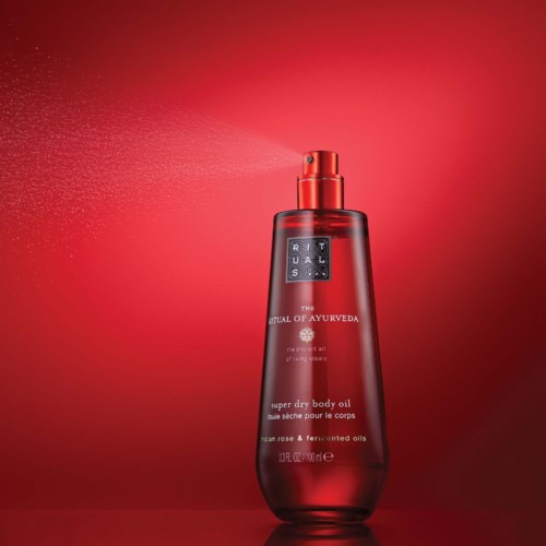 A red body oil spray bottle with a black label and spray nozzle, emitting a fine mist against a red background.