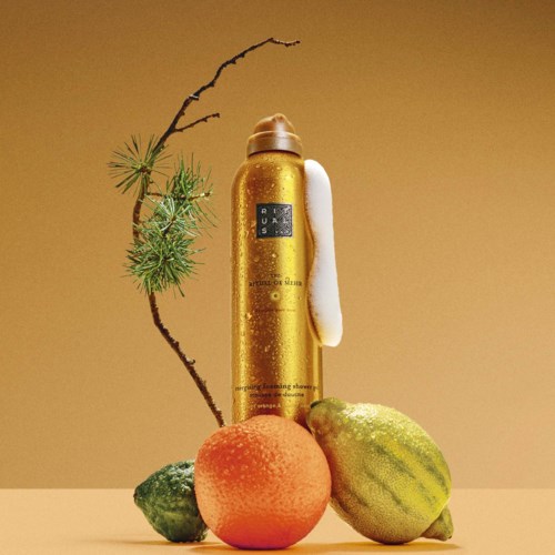 Een gouden fles staat achter een perzik, een kleine groene vrucht en een gestreepte meloen, met een tak en dennennaalden in de buurt.