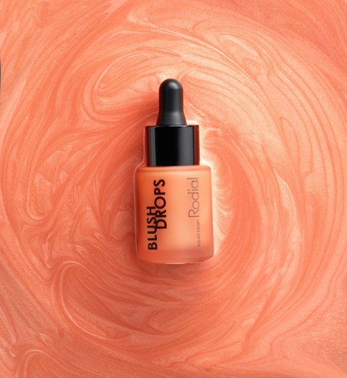 A bottle of blush drops with black cap on a swirling peach-colored background.