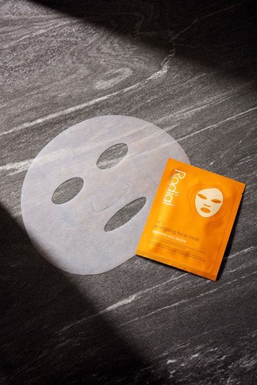 Een gezichtsmasker van papier en een oranje pak liggen op een donkere ondergrond, met licht en schaduw die contrast creëren.