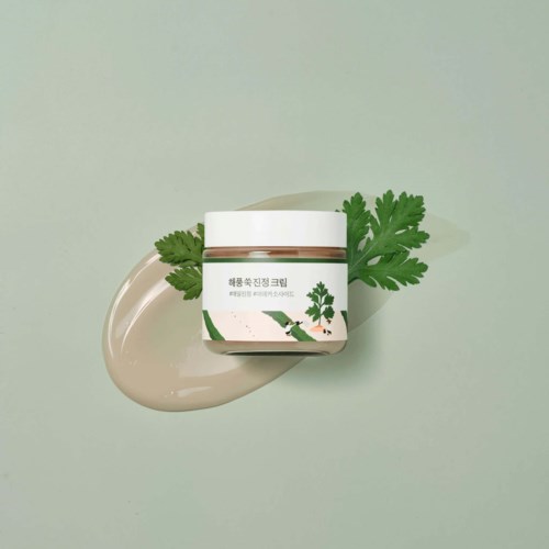 En krukke med creme og grønne blade, placeret på en lys grøn overflade med creme ved siden af.
