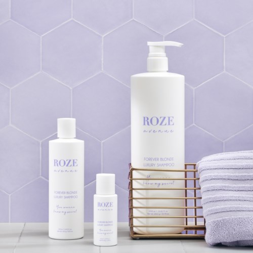Tre hvite flasker med sjampo eller lotion med lavendelskrift, et brettet håndkle og en trådkurv på en lilla flislagt vegg.