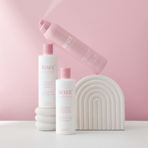 Tre flasker sjampo og balsam med rosa lokk, arrangert på hvite bølgende plattformer mot en lys rosa bakgrunn.