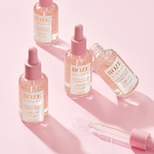 Fyra rosa tonade flaskor med glow tanning drops och pipettkorkar på en rosa bakgrund.