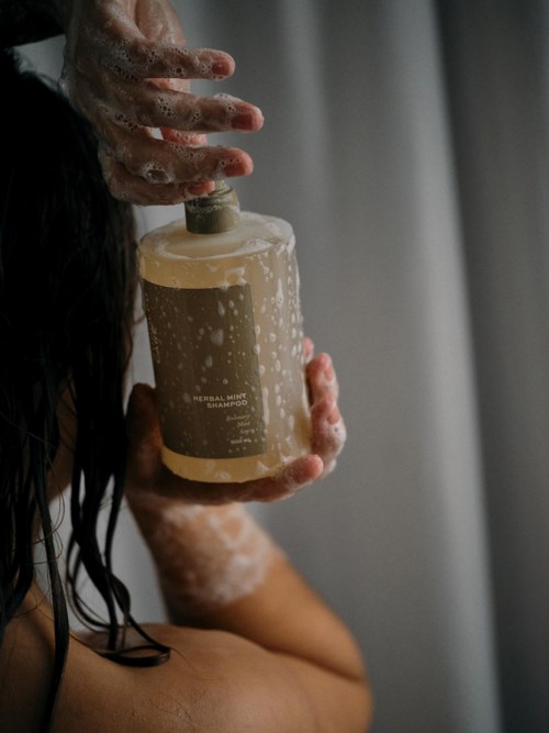 Person vasker hår med shampoo, sæbeskum er synlig, holder en shampoo-flaske.