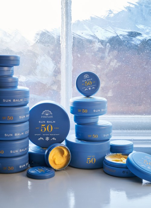 Staplar av blå solskyddsförpackningar med SPF 50 står på ett fönsterbräde. Några lock är öppna, visar gul salva.