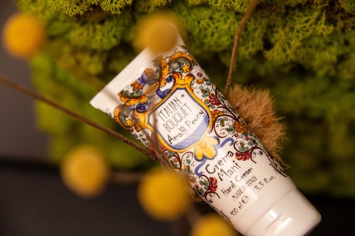 A floral-scented hand cream tube with ornate label, surrounded by greenery and small yellow flowers.