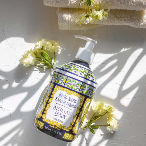 Eine Flasche Flüssigseife mit Sicilia Zitronenduft. Umgeben von weißen Blumen und Handtüchern, mit Schatten auf der Oberfläche.