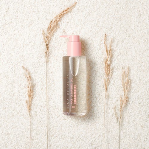 En tydlig flaska med rosa pump och etikett, omgiven av torkat gräs på en texturerad bakgrund.