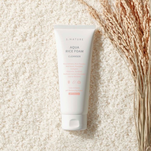 En tub av S.Nature Aqua Rice Foam Cleanser på vita riskorn med torkade växter bredvid.