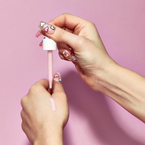 Händer med färgglad nageldekoration med hjärtan som håller ett rosa läppglansborste mot en rosa bakgrund.
