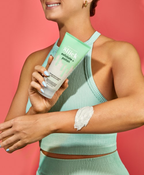 En kvinna i träningskläder applicerar kräm på armen, håller tuben med exfolieringsprodukt, ler.