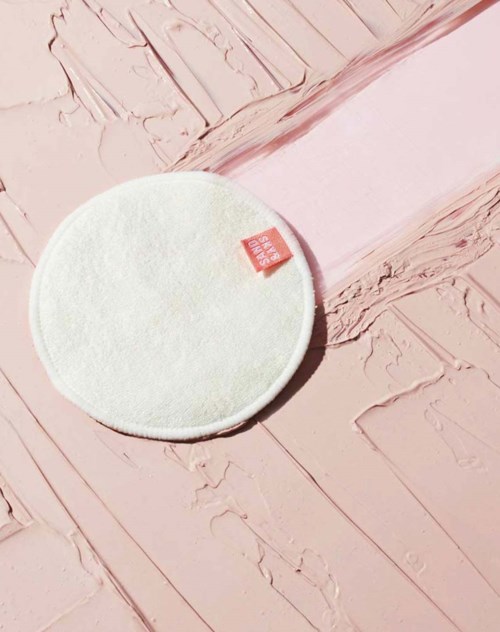 En hvit rund sminksvamp med en liten rød merkelapp på en teksturert rosa plate.