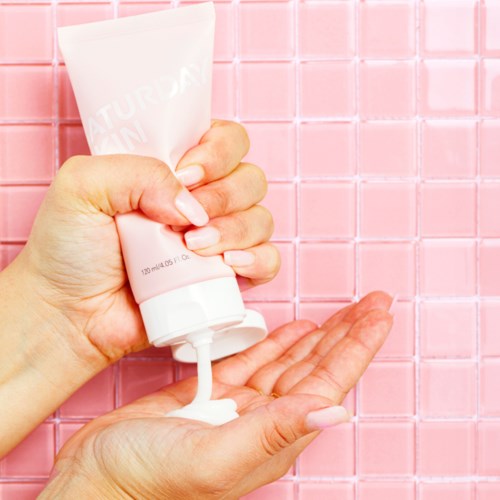 En person som kramar ut vit lotion från ett rör på handflatan mot en rosa kaklad bakgrund.