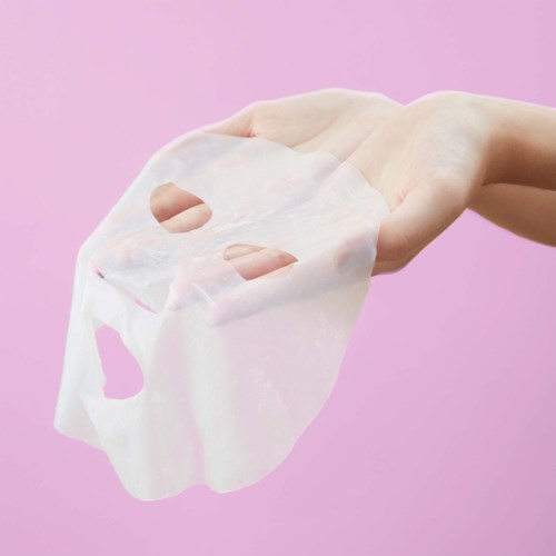 Een hand houdt een wit gezichtsmasker tegen een roze achtergrond.