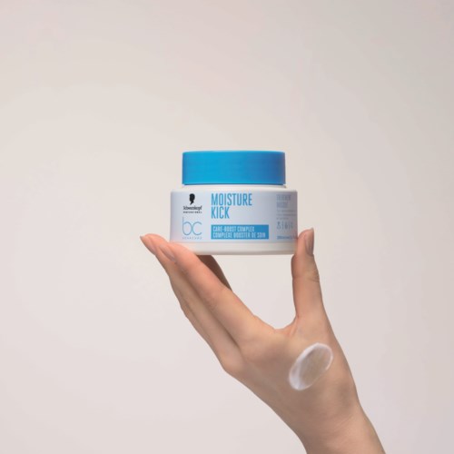 Een hand houdt een blauwe en witte container met het label Moisture Kick, met een beetje crème op de vinger.