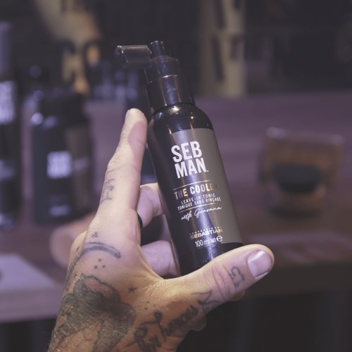 Een hand houdt een zwarte fles met de tekst "SEB MAN THE COOLER" in een zwak verlichte omgeving.