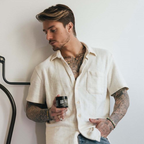 En tatuerad man som håller ett litet burk, står mot en ljus vägg med ett svart föremål nära.