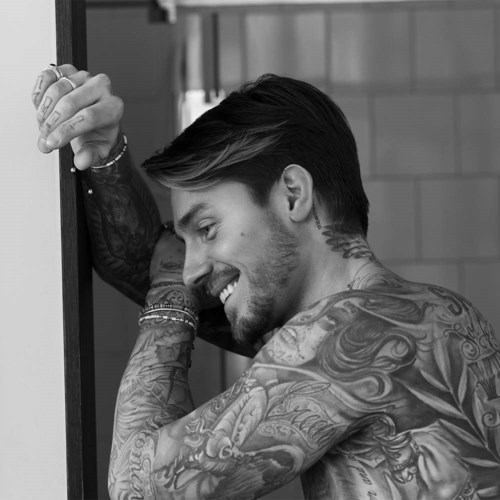 En leende man med tatueringar vilar sitt huvud mot en vägg, visar glädje och kroppskonst.
