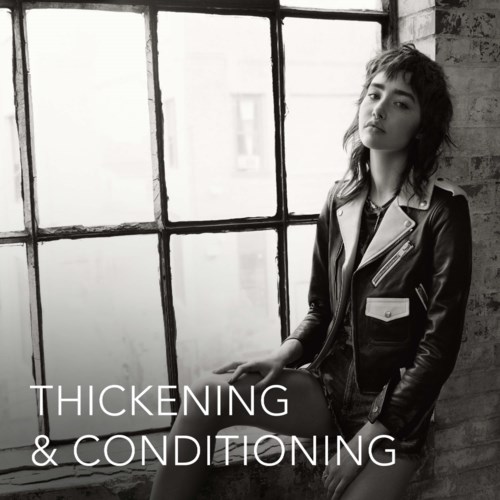 A young woman with wavy hair sits by a large window, wearing a leather jacket in a black-and-white photo.