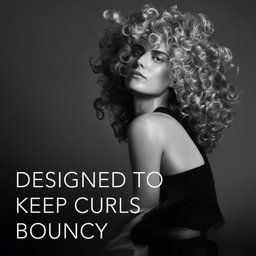 En kvinne med stor, krøllete hår sitter med skulderen synlig, ser selvsikker ut. Tekst sier: Designed to keep curls bouncy.
