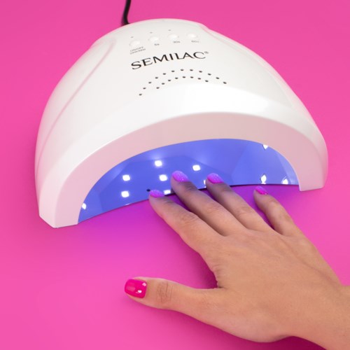 A white UV nail lamp with blue light curing nails, placed on a pink surface, with a person's hand underneath.