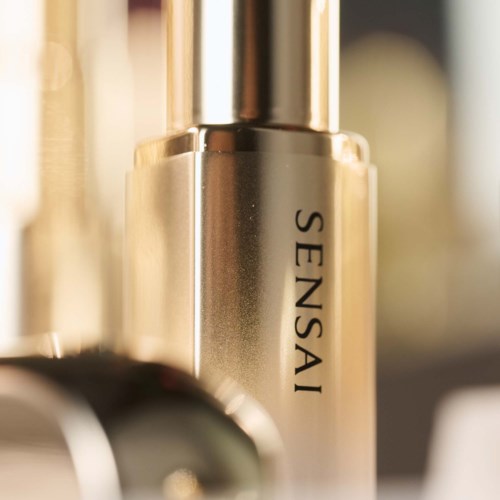 En gylden parfume flaske med navnet Sensai skrevet på, står oprejst på en overflade.