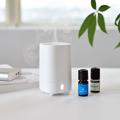 En vit aromdiffusor avger ånga, med två små blå och svarta flaskor bredvid, på en vit yta.