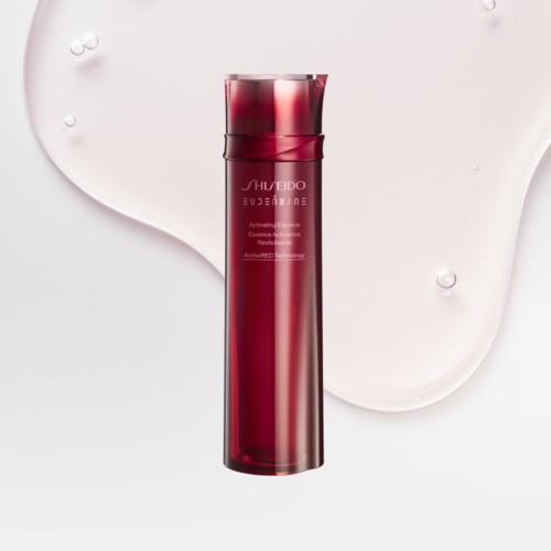Eine hohe, rote Hautpflegeflasche mit elegantem Design auf weißem Hintergrund, in der Nähe ist etwas Flüssigkeit verschüttet.