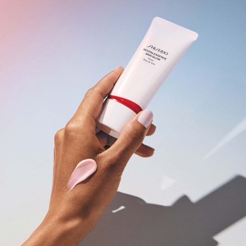 En hand håller en vit tub med hudvårdsprodukt med ett rött band, med en lätt makeupkräm på fingret. Enkel bakgrund.