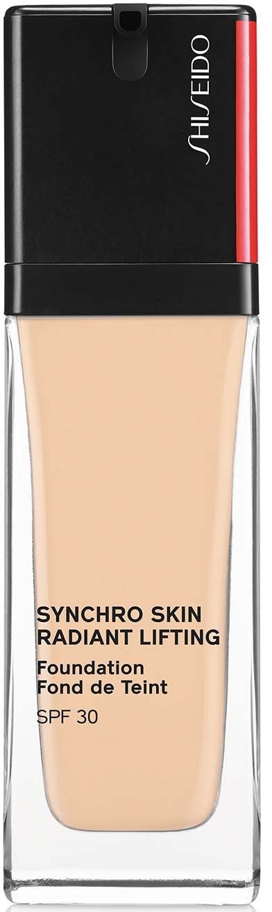 En fondationflaska med svart och röd topp, klart glas, och en beige vätska inuti. Etiketten säger 'SYNCHRO SKIN RADIANT LIFTING'.