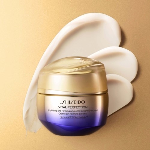 En burk Shiseido Vital Perfection-kräm omgiven av vita krämduttar på en beige bakgrund.