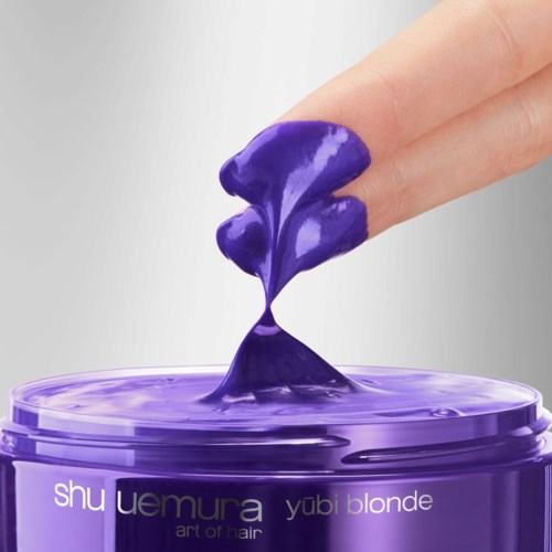 A finger dips into purple hair wax, showing its creamy texture above a jar. The jar is purple with white and black text.