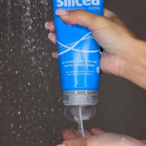 Hænder der hælder shampoo fra en blå flaske i håndfladen over en grå baggrund.
