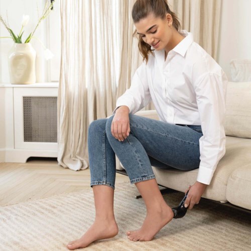 A woman in a white shirt and jeans is sitting on a sofa, putting on black slippers.