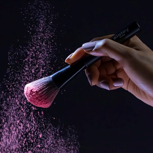 En hand skakar en makeup-borste, rosa pulver sprids i luften mot en mörk bakgrund.