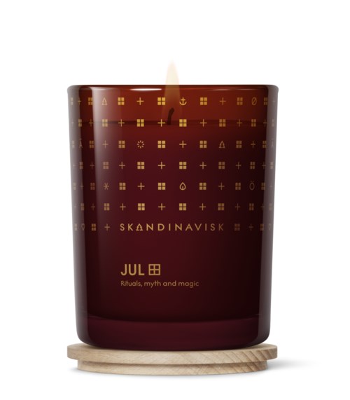 A red candle in a jar with golden symbols and text, lit and on a wooden coaster.