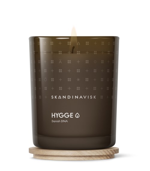A brown candle in a jar, labeled 'SKANDINAVISK HYGGÉ Danish DNA,' on a small wooden coaster, with a small flame.