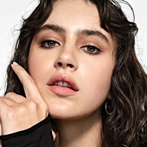 A woman with wavy hair and makeup touches her face, looking confident and relaxed.