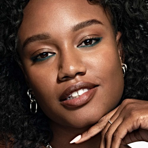A woman with curly hair, wearing earrings, has natural makeup and touches her face gently.