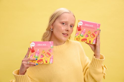 En ung kvinna med blont hår i en gul tröja håller två rosa mini kit-boxar framför en gul bakgrund.