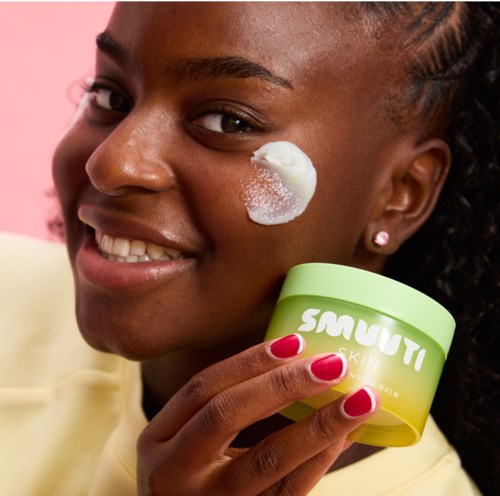 Een vrouw glimlacht terwijl ze crème op haar wang aanbrengt en een groene container met Smuti Skin houdt.